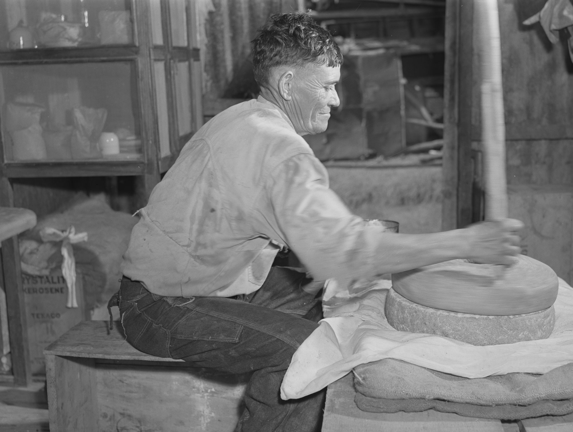 A photograph of a man using grinding stones on St. Croix Island in 1941. The man is sitting on a wooden box or stool, in front of a wood-plank table. He is wearing a long-sleeved shirt and what appear to be jeans. On the table are two large circular stones, one stacked on top of the other. The man is using a wooden pole placed in a depression near the edge of the upper stone to spin in on top of the lower stone, grinding maize grains between them.