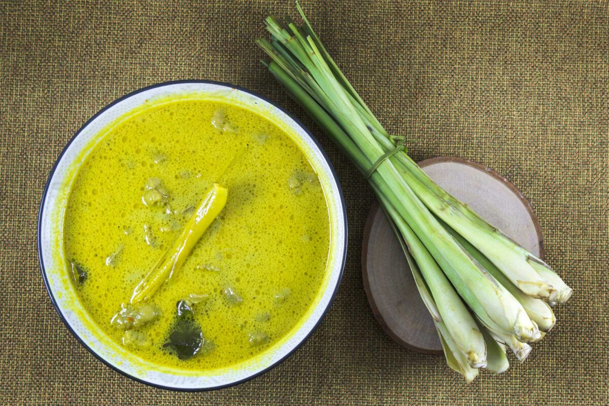 Photograph showing a bowl of food (apparently curry, it is a yellow liquid with chunks of plants floating in it) with a bundle of lemongrass on a slice of wood next to it. 