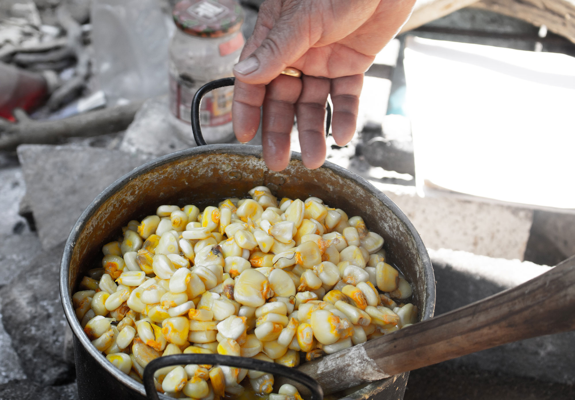 Photograph of nixtamalization of maize. The photo shows a metal pot with two handles that is filled with corn kernels. A person, mostly out of frame, is stirring the maize with a wooden spoon. The spoon has a white stain on it near the level of the pot, perhaps caused by the added lime. Cement blocks, plastic bottles, and other debris can be seen in the background.