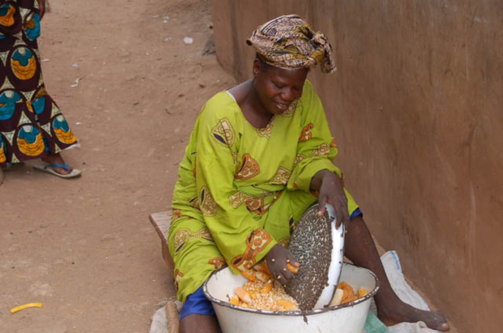 A woman shelling maize in Nigeria. The woman is sitting on a wooden stool near a brown wall using a maize sheller to remove maize grains from maize ears and catch the grains in a large white metal bowl. The sheller looks like a round disk with bumps on the surface, and the woman appears to be rubbing an ear of corn over it. The woman is wearing a kerchief on her head, a long, green tunic, and blue pants that stop just below the knee.