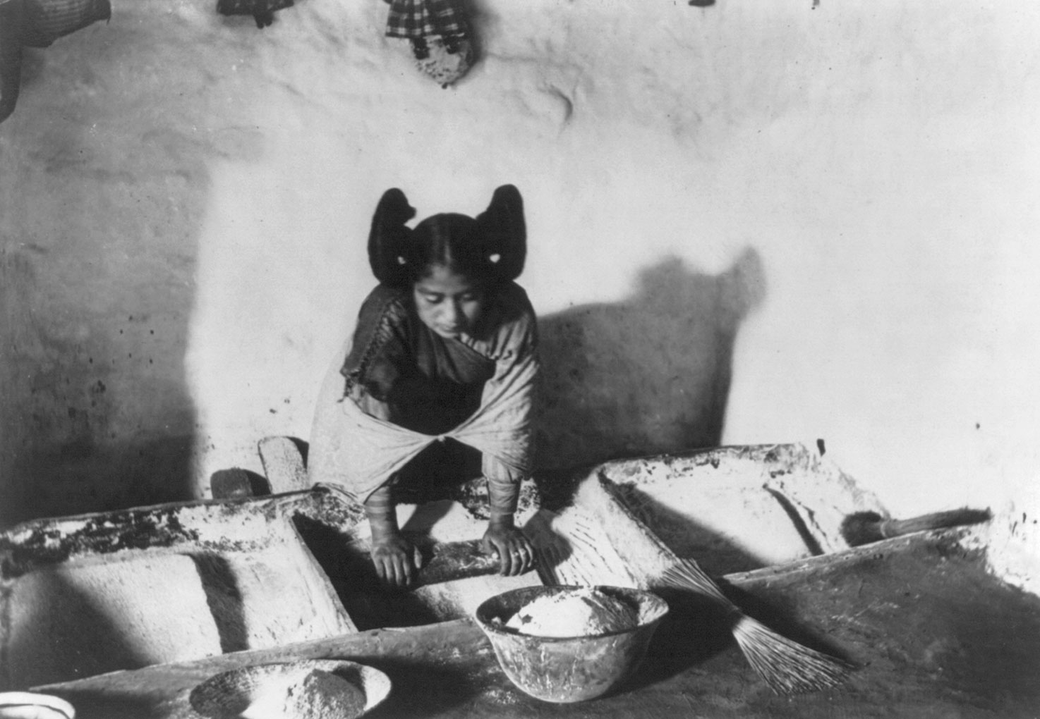 Black and white photo of a young Hopi woman grinding corn taken around 1909. The photo shows a young woman wearing a shirt or dress and a shawl. Her hair is in a squash-blossom hairstyle, with one bun on each side of her head. She is using both hands to press down on an oval stone, using it to grind maize. In front of her sit bowls of maize flour and a whisk broom.