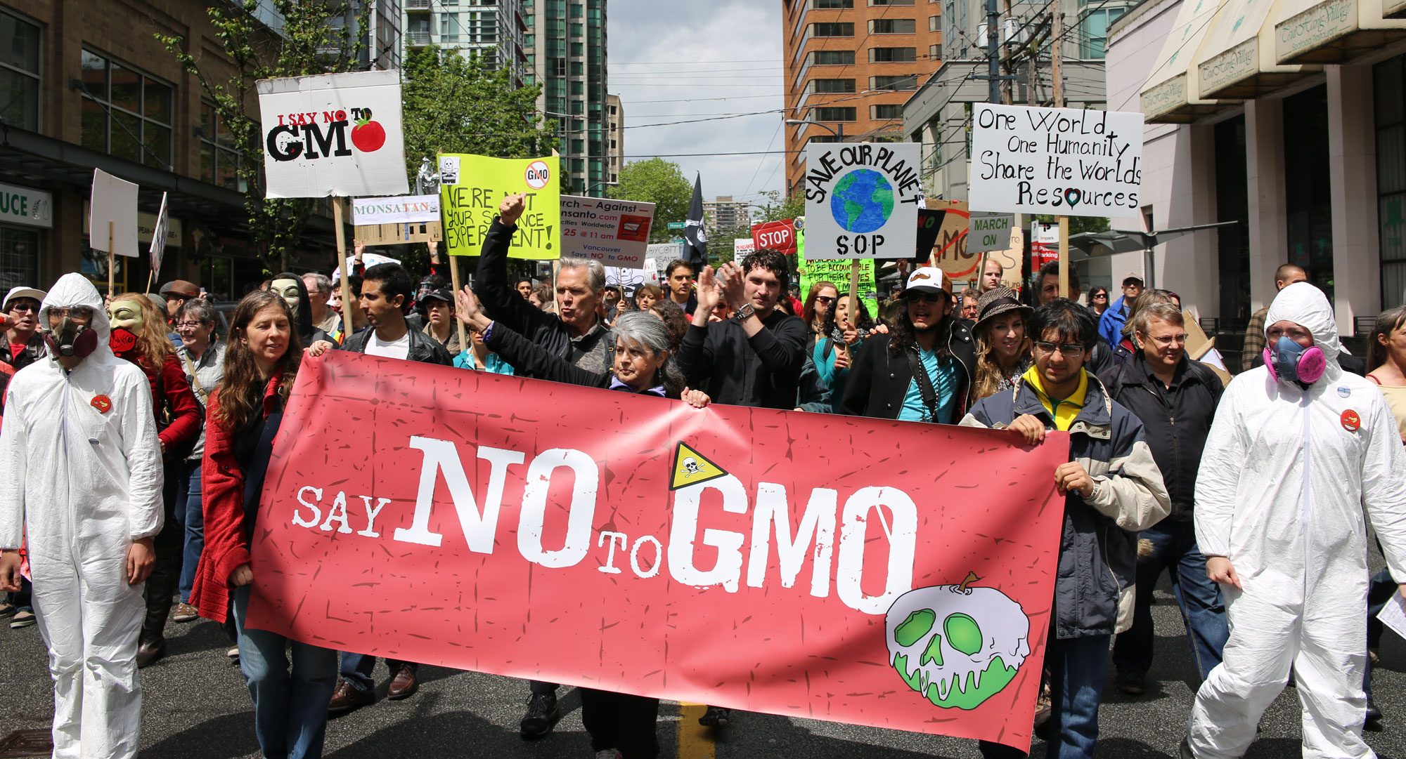 Photograph of a protest march against Monsanto. The photo shows a group of people marching down a street between buildings. The closest people hold a red banner that says "Say no to GMO" on it. The banner is flanked by two people in white hazmat suits and respirators. Other protest signs say things like "I say no to GMO," "Save our planet SOP," and "One world one humanity share the world's resources."