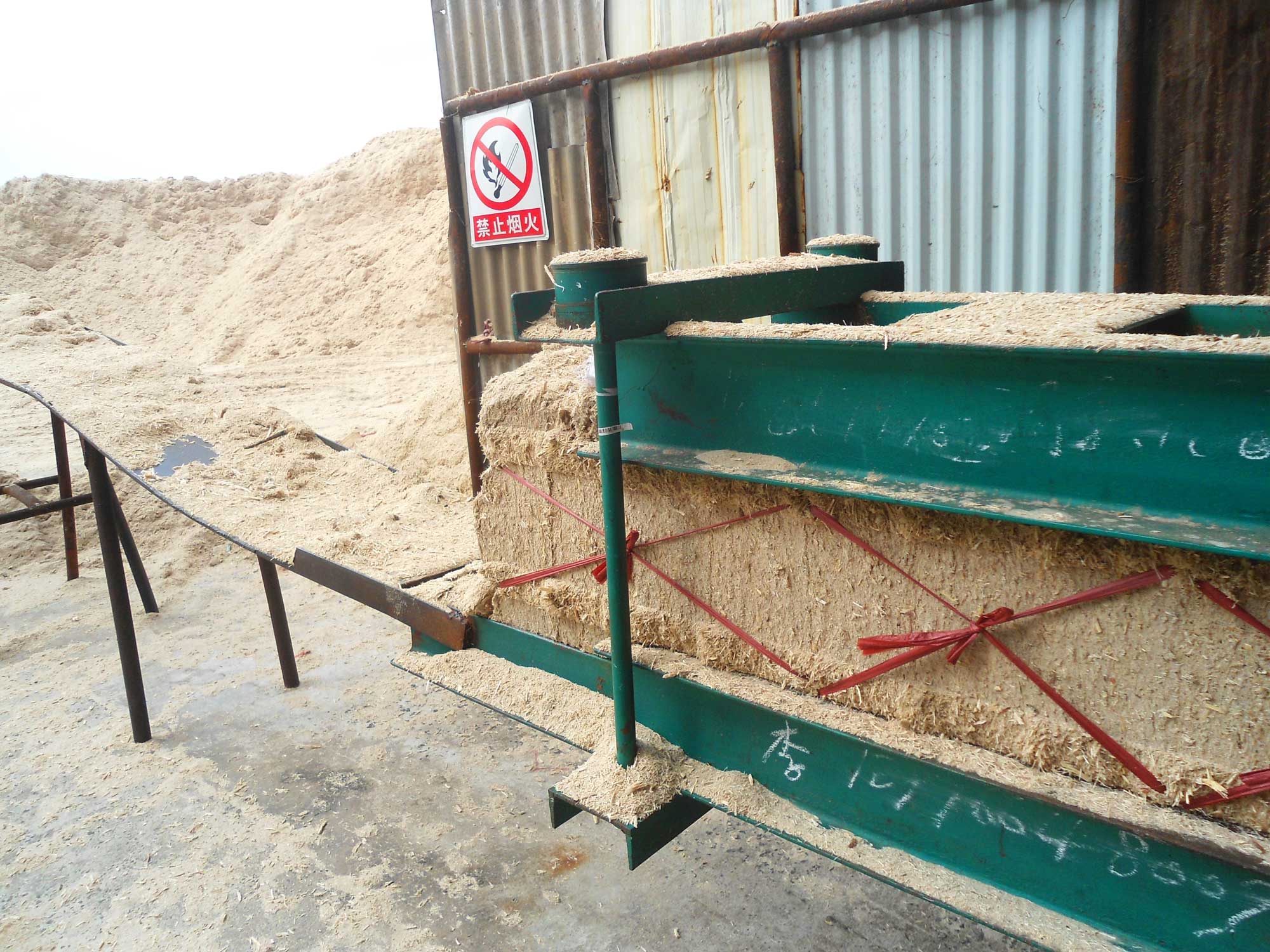 Photograph of a sugarcane bagasse bailing machine in Hainan, China. The photo shows beige fibrous material packed into rectangular blocks and tied with red plastic emerging from a chute. Piles of bagasse can be seen in the background.
