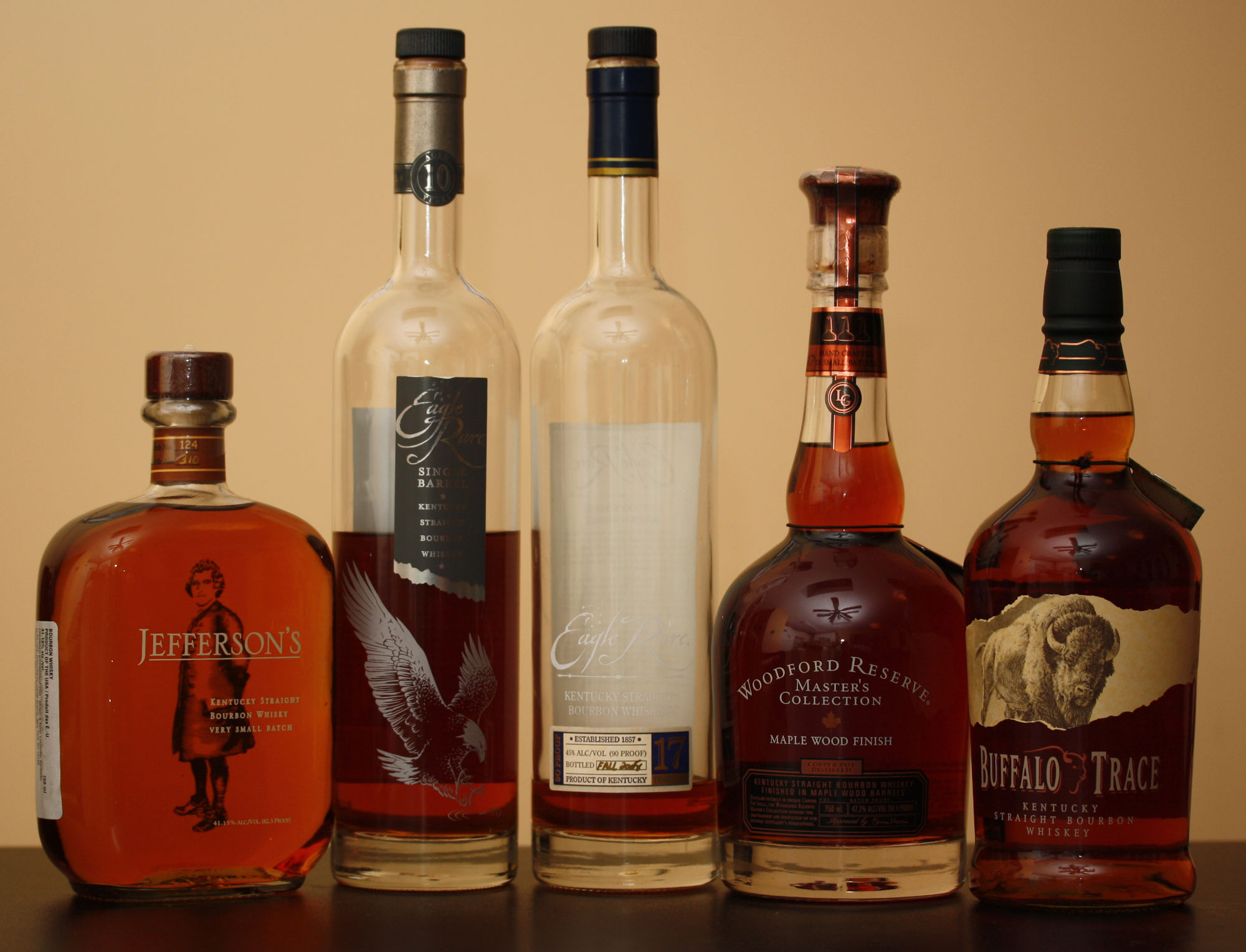 Photograph of a row of bottles of bourbon. The photo shows five bottles of bourbon of a range of shapes and heights, with varying amounts of bourbon left in them. The bourbon is orange-brown in color.