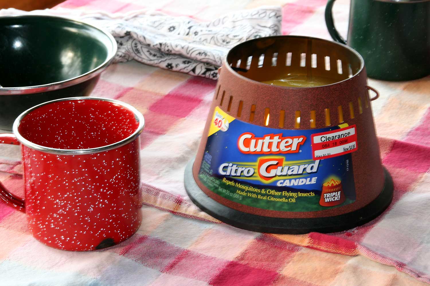 Photograph of a cintronella candle sitting on a gingham table cloth near metal dishware. The candle is in a tapering container with a label on the side that says "Cutter Citro Guard Candle, Repels mosquitoes & other flying insects made with real citronella oil."