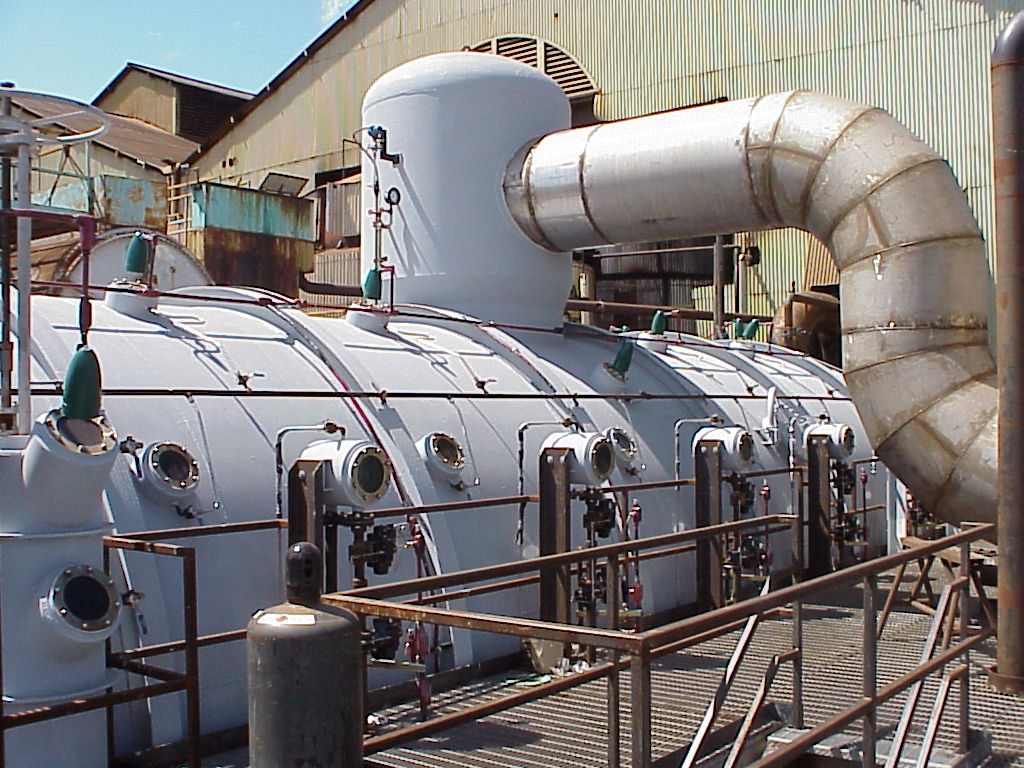 Photograph of a continuous vacuum pan in Maui, Hawaii. The pan is a white cylindrical structural resting on its long side with ports. A duct emerges from a smaller white cylinder on top of the vacuum pan. The apparatus looks like a submarine.