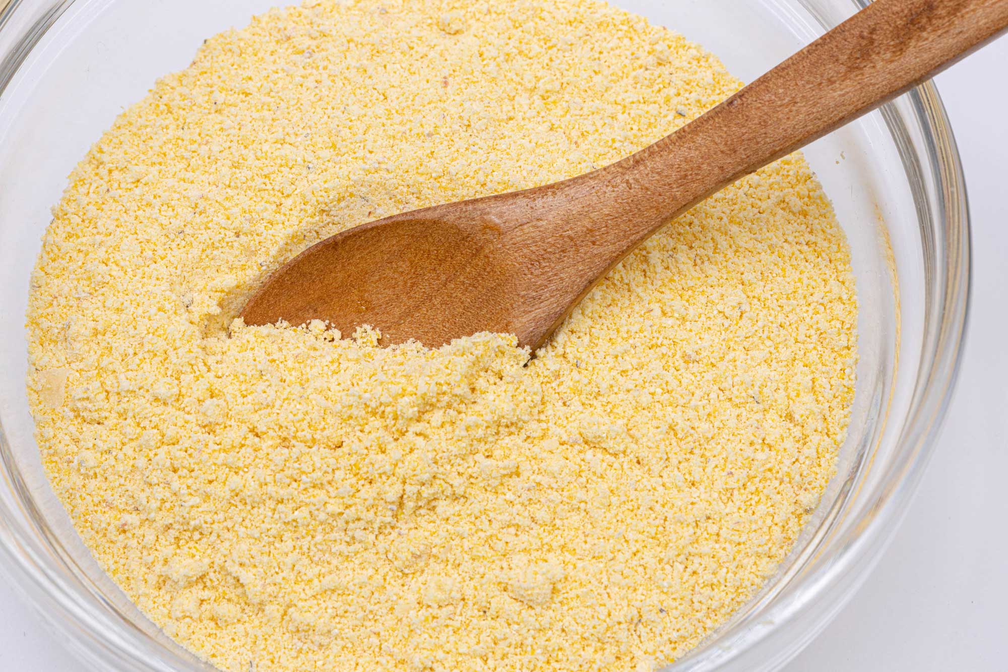 Photograph of corn flour. The photo shows a clear glass bowl full of corn flour. The flour is light yellow in color and looks granular. A wooden spoon is resting in the flour.