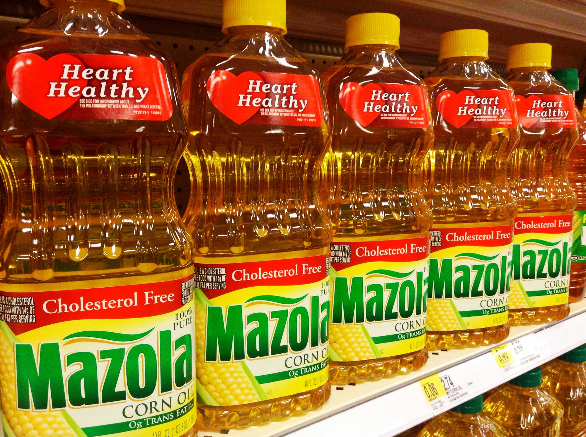 Photograph of bottle of corn oil on a shelf in a store. The photo shows a row of Mazola brand corn oil in clear plastic bottles with yellow and green labels. Each bottle has a red label that says "Heart Healthy" and a red banner on the brand label that says "Cholesterol Free."