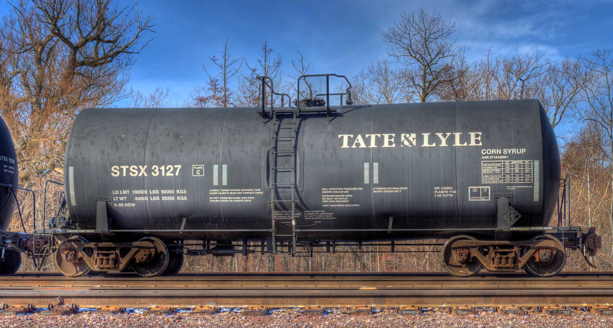 Photograph of a black tanker railcar sitting on a railroad track. The side of the car has lettering, including the label "corn syrup."