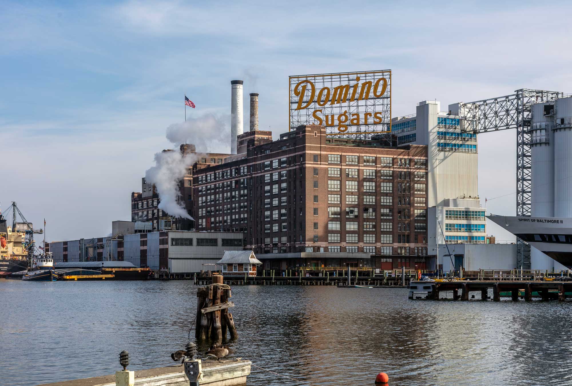 Photograph of the Domino Sugar Refinery in Baltimore, Maryland, U.S.A. The photo shows a brick building with may windows. A large sign that says "Domino Sugars" is on the roof of the building, and white smoke or steam is emerging from the side. Two smokestacks can be seen in the background. The building is sitting at the edge of a body of water.
