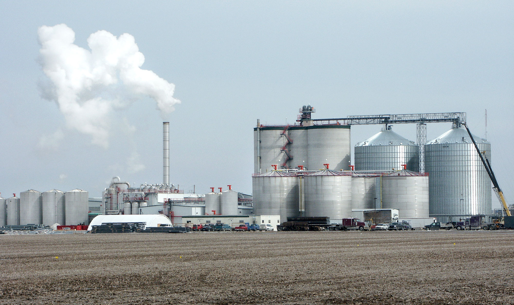 Photograph of a bioethanol plant standing in a barren field. The plant consists of a cluster of cylindrical towers and low buildings. White smoke or steam rises from a thin smokestack at the plant.