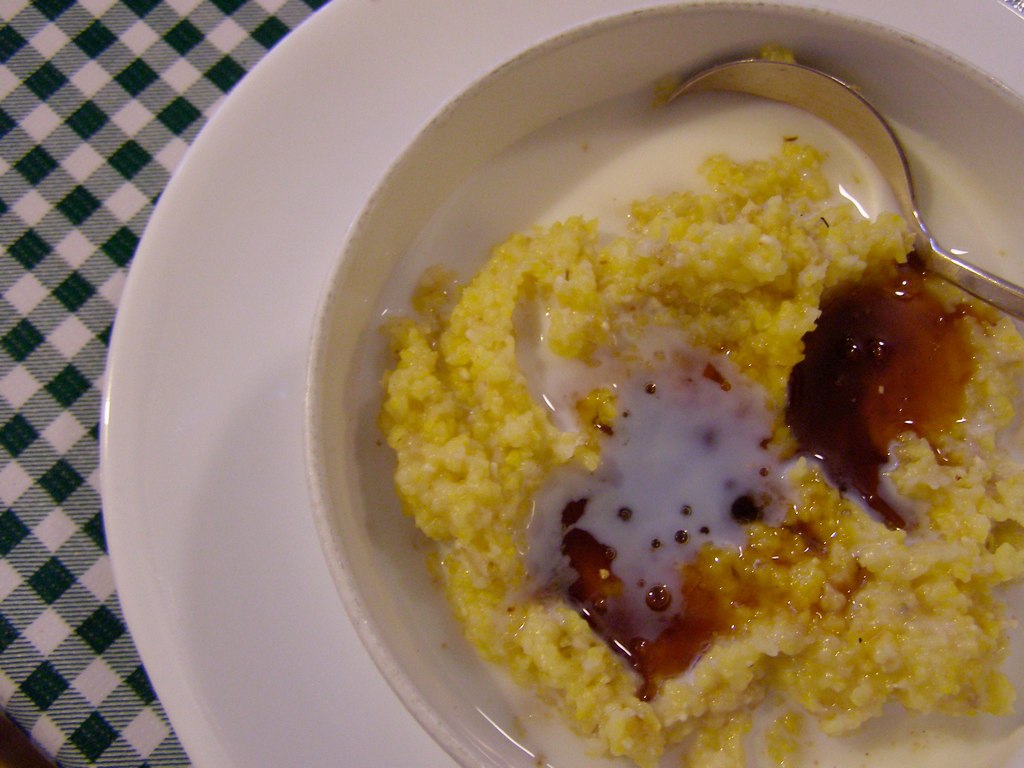 Photograph of grits and milk with sorghum syrup. The photo shows yellow grits (coarsely ground corn) in milk, with brownish syrup on top, all in a white bowl.