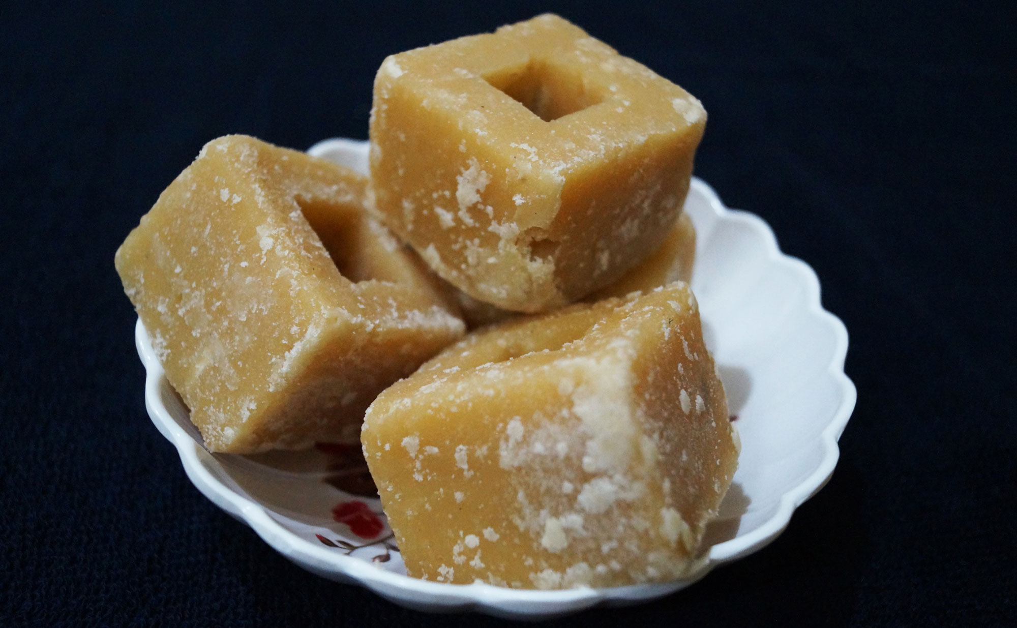 Photograph of jaggery cubes. The photo shows three caramel-colored square cubes with hollow centers sitting on a white dish with a scalloped edge.