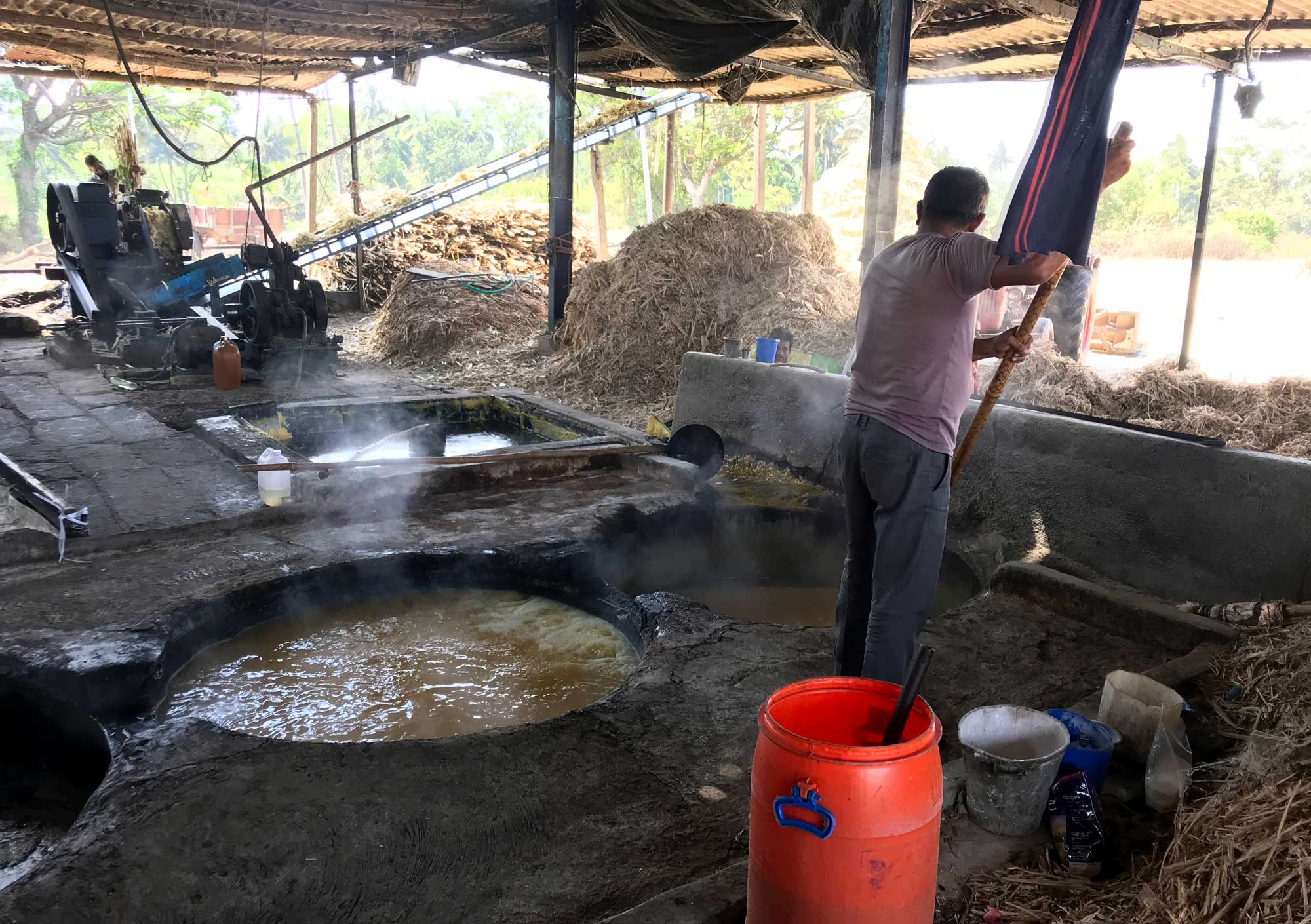 Photograph of a man making jaggery in Karnataka, India. The photo shows a man stirring cane syrup cooking in a pan in the ground with a long pole. Another pan with cooking syrup is on his left. In the background, a grinding mill and large piles of bagasse can be seen.