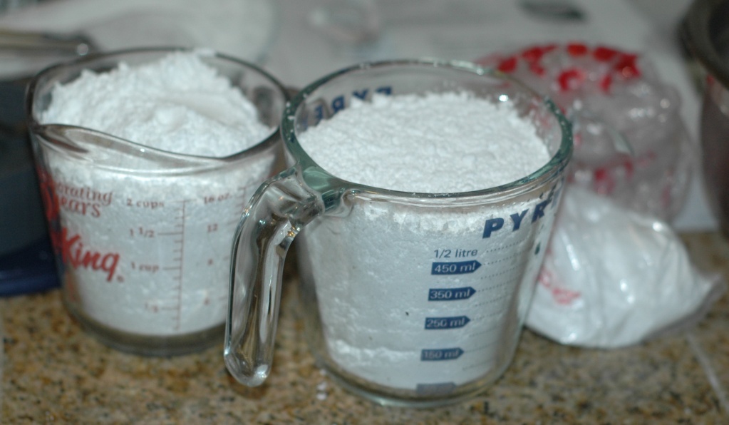 Photograph of powdered sugar or confectioner's sugar. The photo shows two clear glass measuring cups full of a powdery white sugar. A clear bag of powered sugar sits behind them.