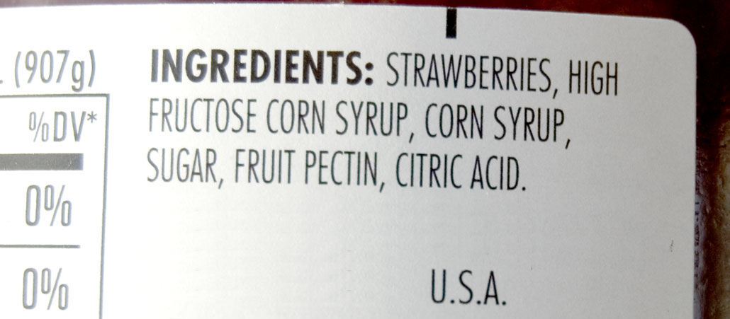 Photograph of an ingredients label for strawberry jam that was made in the U.S.A. Ingredients are strawberries, high fructose corn syrup, corn syrup, sugar, fruit pectin, and citric acid.