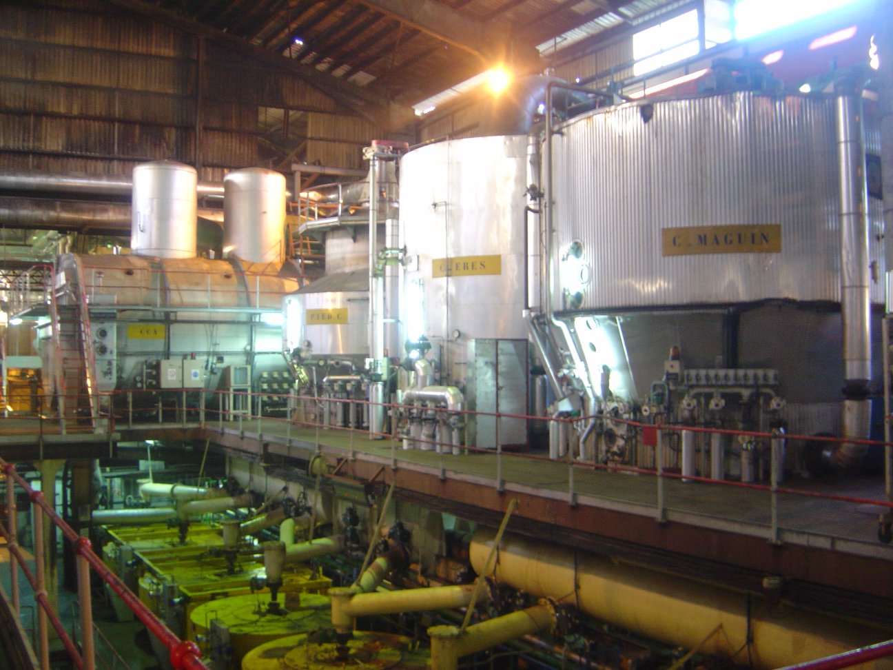 Photograph of sugar vacuum pans and centrifugals in a sugar factory on Réunion island. The photo shows the interior of a plant with cylindrical silver vacuum pans and a row of centrifugals that are on a floor below them. The centrifugals are somewhat difficult to see.