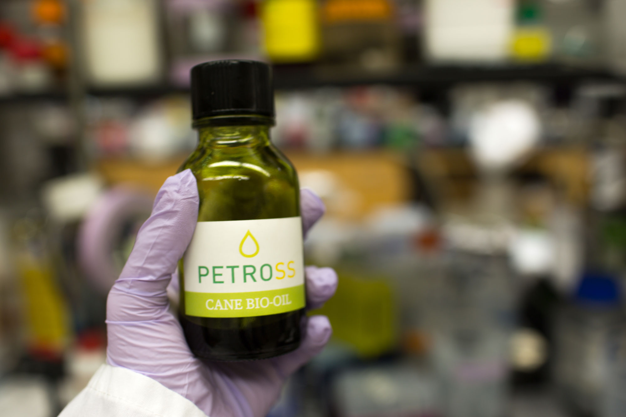 Photograph of gloved hand holding a small bottle made of green glass with a black top. The bottle is labeled "PETROSS CANE BIO-OIL."