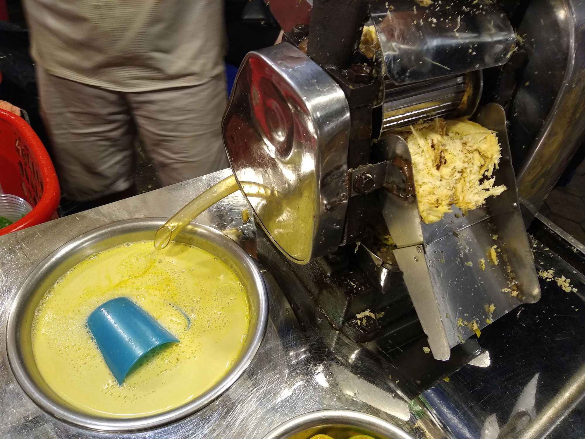 Photograph of fresh sugarcane juice is a stainless steel bowl at a night market in Cambodia. The photo shows an opaque yellowish liquid in a bowl with a blue mug sitting on its side in the liquid. A grinder is next to the bowl of liquid.