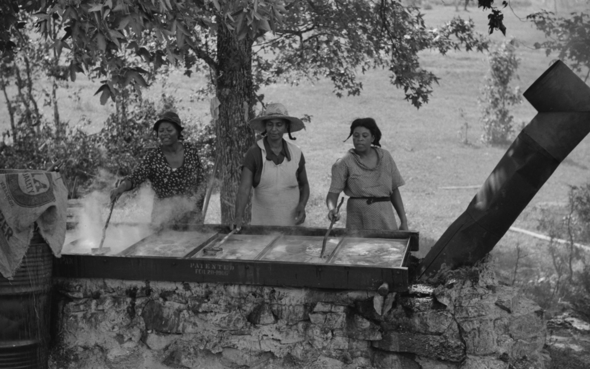 Black and white photograph of three women using ladles to skim the scum off cooking sorghum juice. The juice is cooking in shallow rectangular pan separated into five or six compartments by partitions.