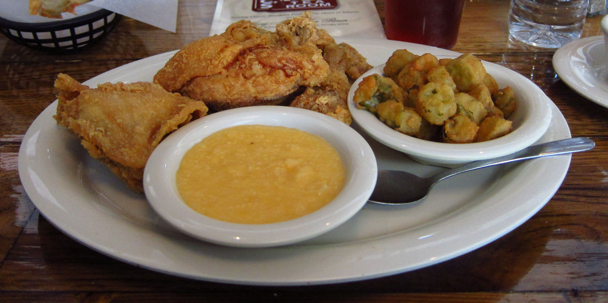 Photograph of a meal at a restaurant in Atlanta, Georgia, U.S.A. The photo shows a round white plate with pieces of breaded, fried chicken, a bowl of breaded, fried okra, and a bowl of grits sitting on it. 