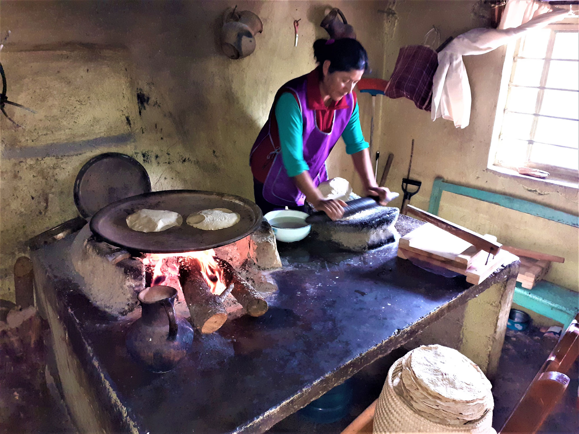 Photograph of a woman preparing tortillas in Oaxaca Mexico. The photo shows a woman using a metate and mano to grind maize and make masa. Next to her is a flat metal sheet sitting a a fire. Two circular tortillas are cooking on the sheet. In from of the table where the woman is working, a basket holds a stack of finished tortillas.