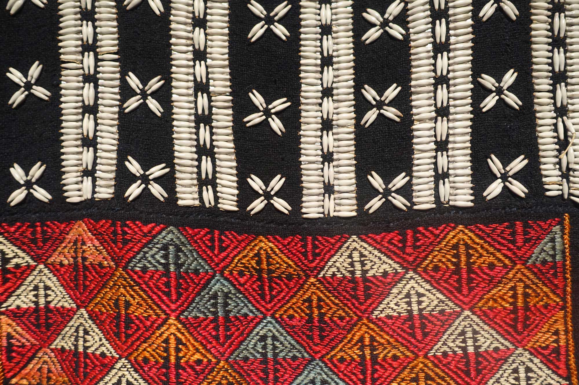 Photograph of a textile decorated with beads from wild adlay, Vietnam. The photo shows part of a textile. The upper part is black whereas the bottom part is multicolored with a woven design. Spindle-shaped adlay beads are organized in designs on the black part of the textile, including "x's" and parallel rows.