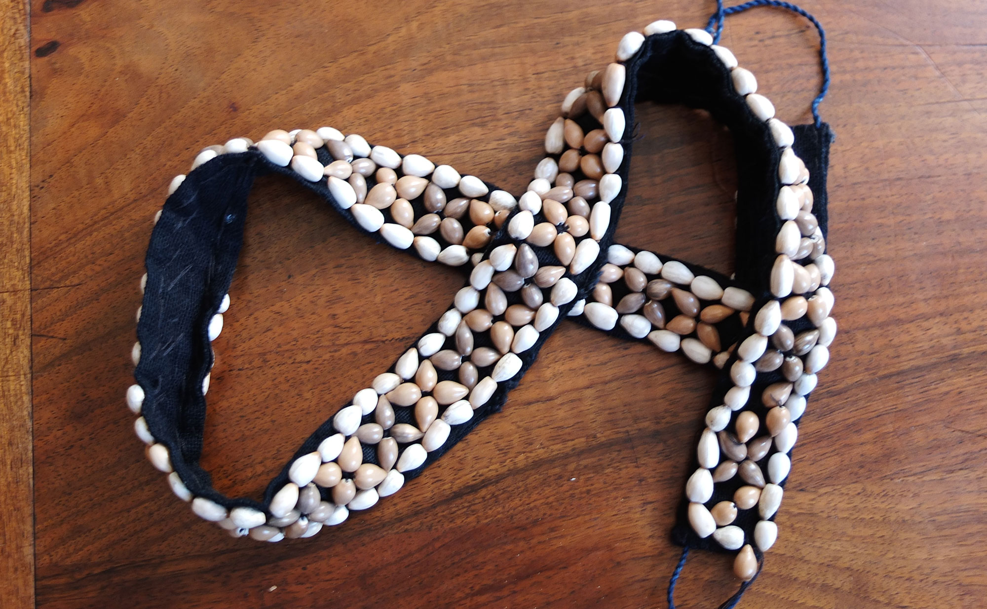 Photograph of a belt decorated with adlay beads made by a member of the Hmong ethic group in Laos. The photo shows a blue fabric belt that has one side decorated with adlay beads. The beads border the belt and are organized in a row of "x's" along the front side of the belt.