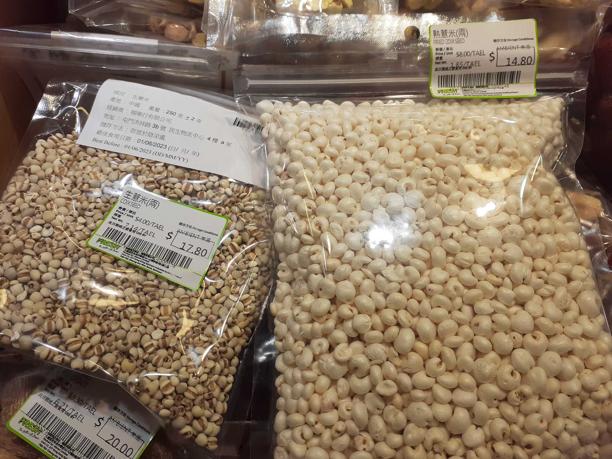 Photograph of bags of adlay grains at a market in Hong Kong. The photo shows two clear plastic bags filled with grain. The bag on the left has smaller, beige grains and is labeled as "coix seed." The bag on the right has larger white grains and is labeled as "fried coix seed."