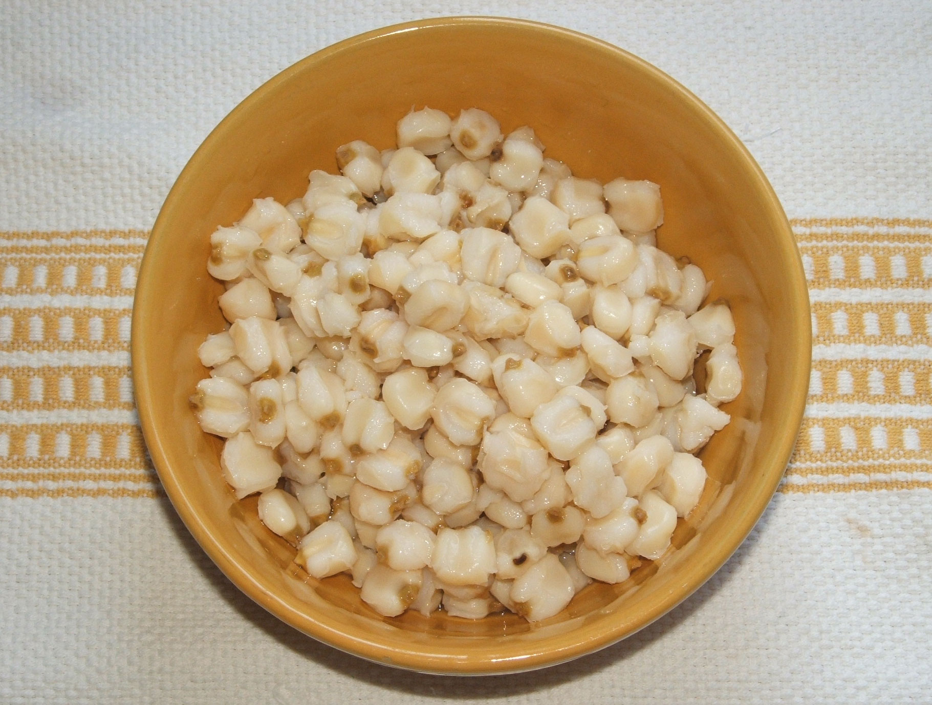 Photograph of hominy. The photo shows hominy, wet, white maize kernels, in a tan bowl sitting on a white cloth with a yellow-striped pattern on it. 