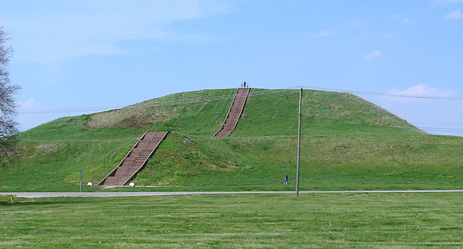 Photograph of Monks Mound at the Cahokia site in Illinois, U.S.A. The photo shows a large earthen mound with two tiers covered with grass. Steps run straight from the base to the top of the mound.