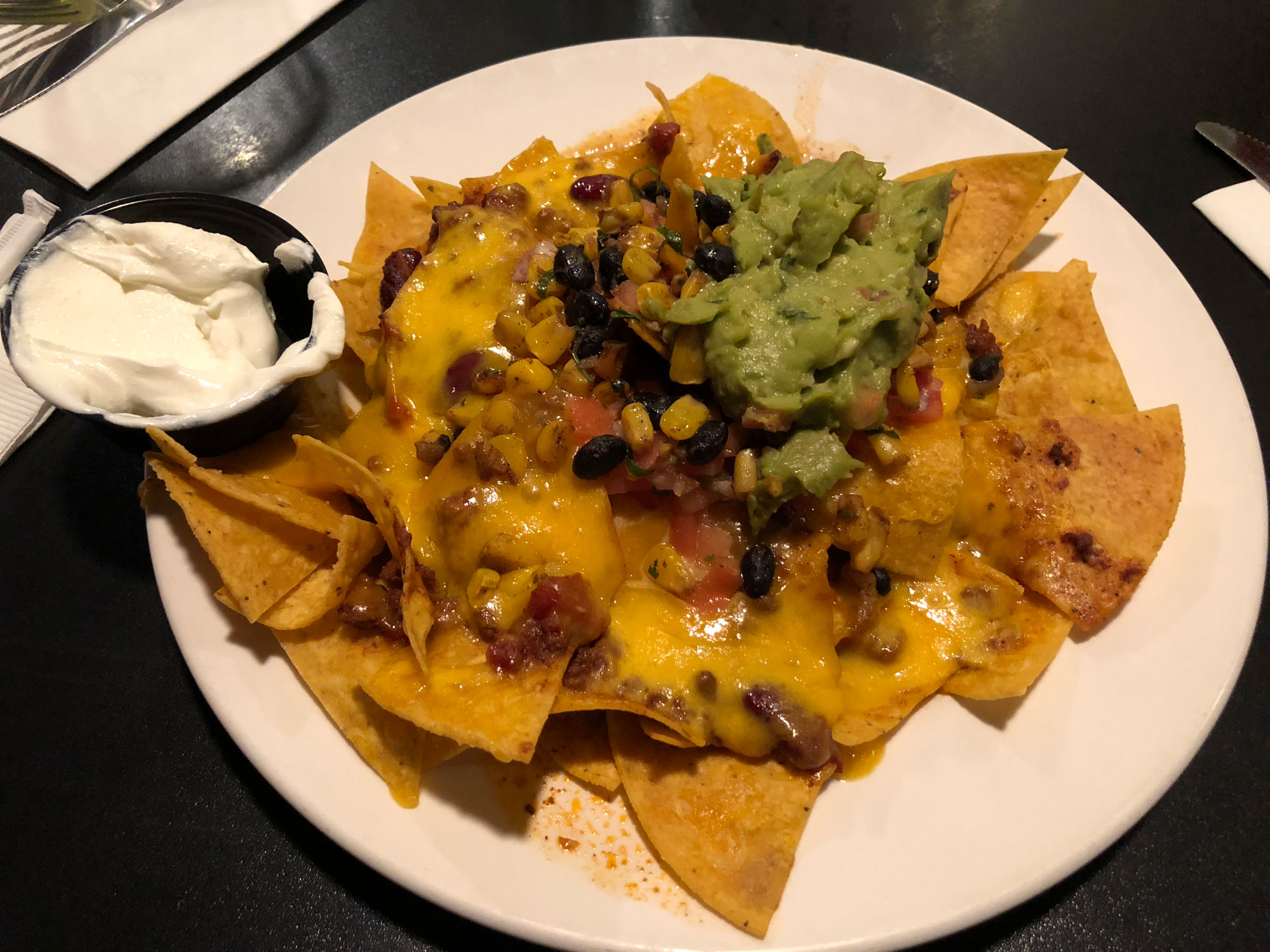 Photograph of a plate of nachos. The photo shows a round white plate on a black surface. On the plate are tortilla chips covered with melted cheese, meat, corn kernels, black beans, and guacamole. A small black container of sour cream is on the edge of the plate next to the chips.