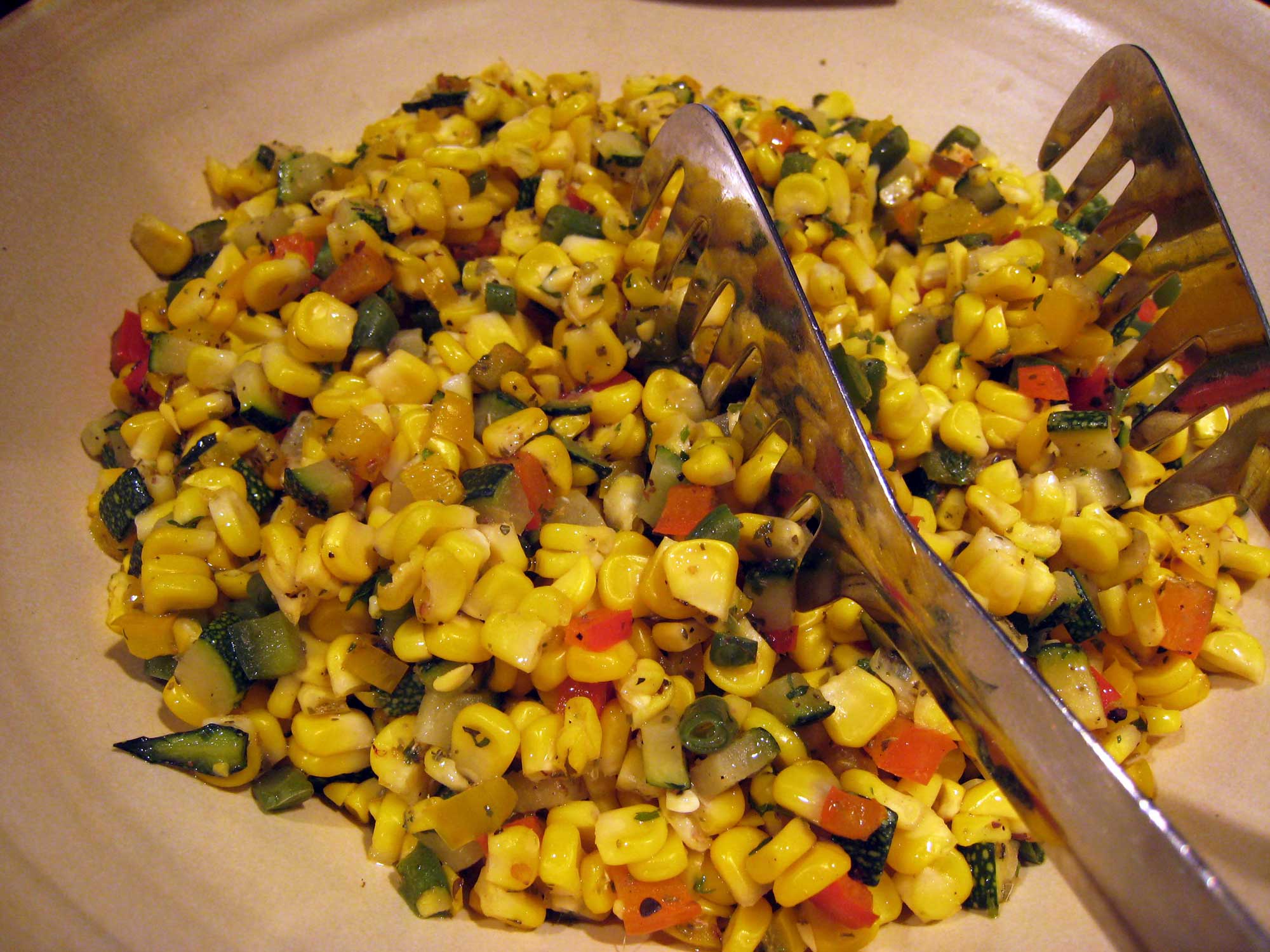 Photograph of succotash. The photo shows a mixture of cooked maize kernels with other ingredients in a white bowl with metal tongs resting in it.