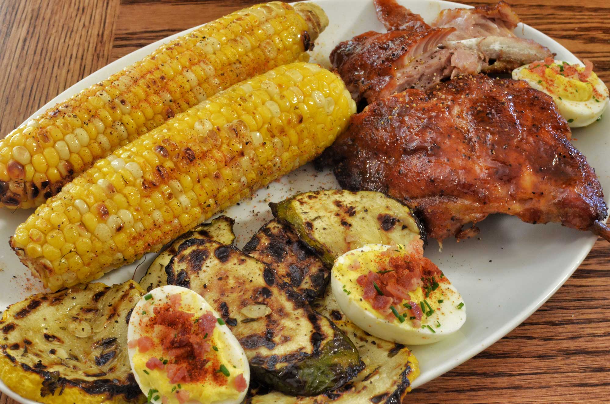 Photograph of a meal including sweet corn on the cob. The photo shows ribs, grilled squash slices, deviled eggs, and two sweet corn cobs on a white plate sitting on a wooden surface.
