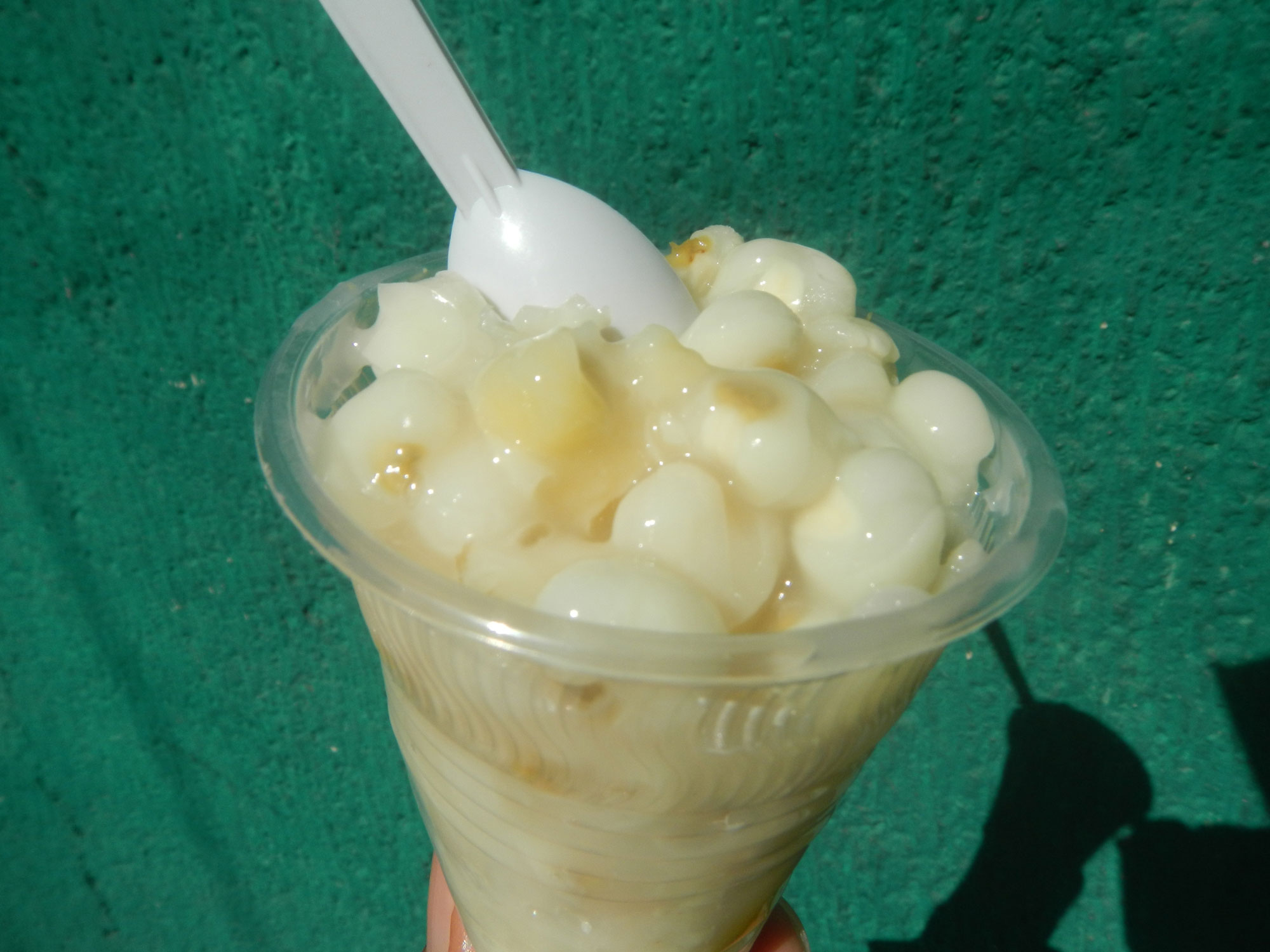 Photograph of binatog, a maize-based dessert from the Philippines. The photo shows a clear, plastic, conical cup filled with white boiled maize kernels with a white plastic spoon standing upright in the kernels. 