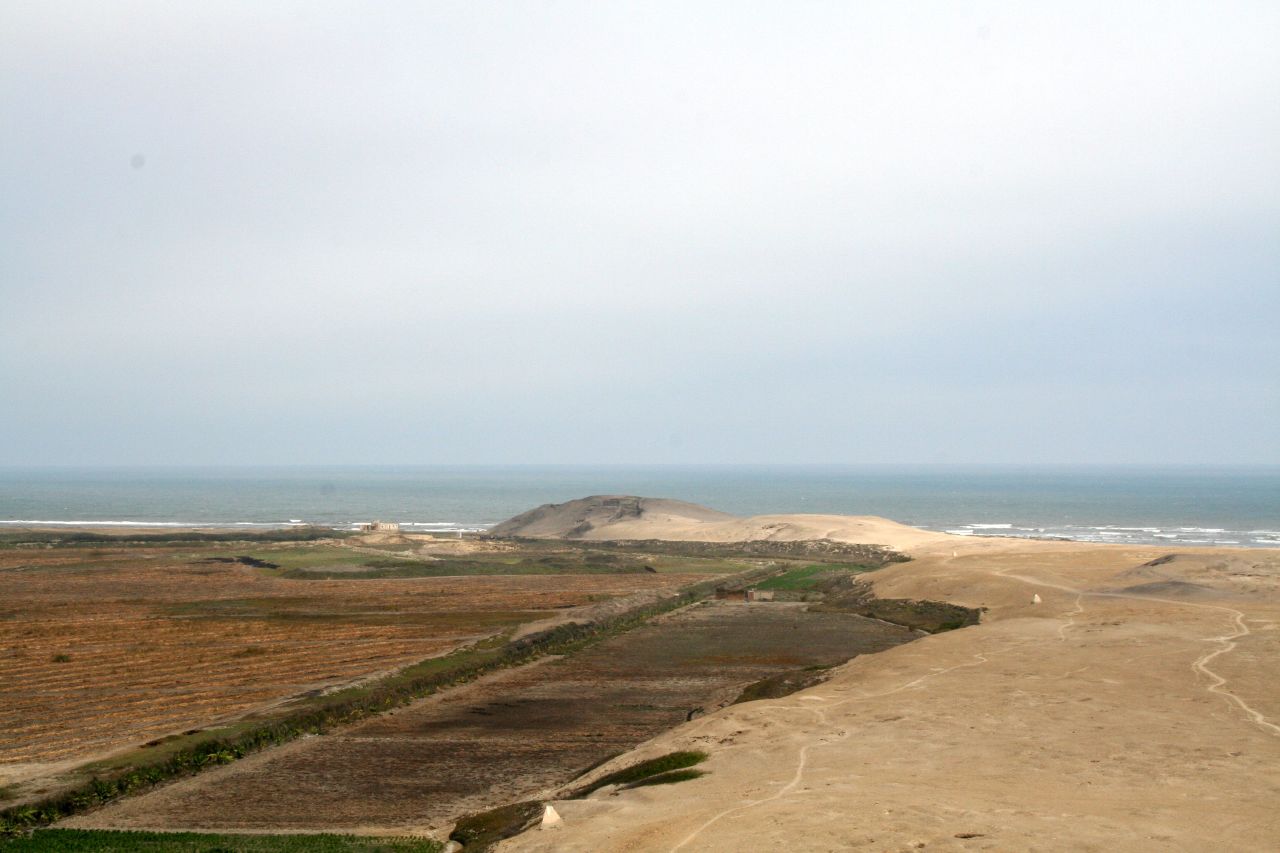 Photograph of the Huaca Prieta site on the coast of Peru. The photo shows a dry landscape with a low hill in the background next to the ocean.