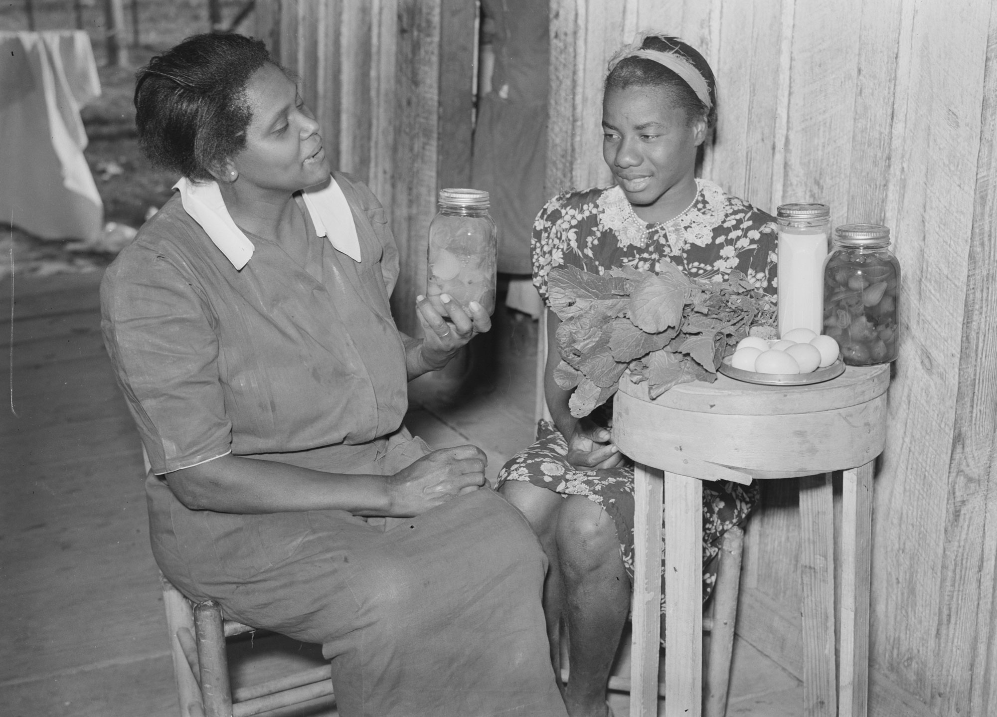 Black-and-white photograph of two women sitting at a small table. The woman on the left sits in a chair and holds of a jar of food while speaking to the other woman. The woman on the right sits on a chair placed against a wall and behind the small table. Greens, eggs, a jar of milk, and another jar of food sit on the table.
