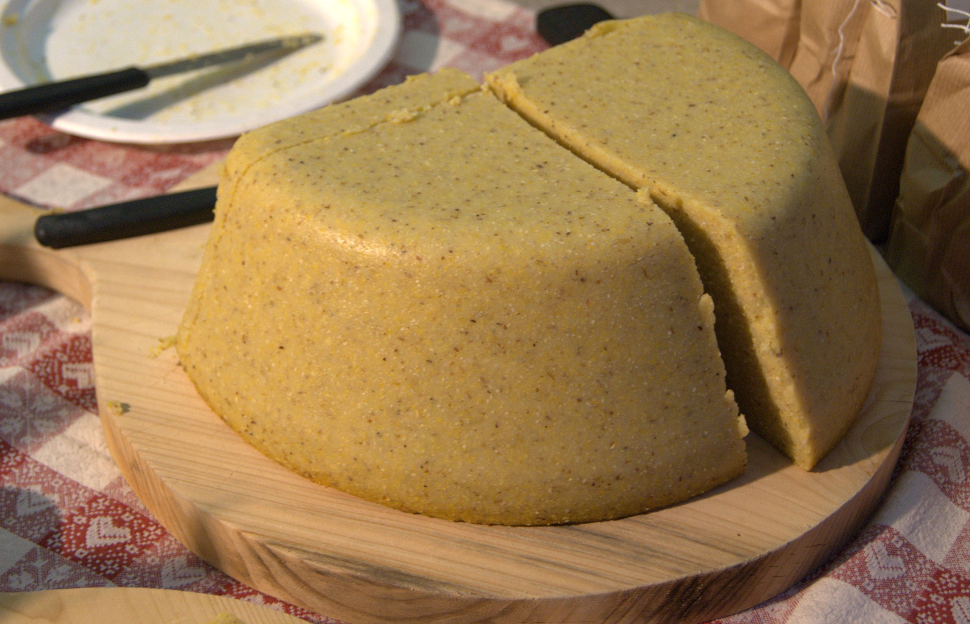 Photograph of polenta. The photo sows half of a round cake of polenta sitting on a wooden cutting board. The polenta has been cut into two halves, with a thinner slice also cut from the side not facing the viewer.