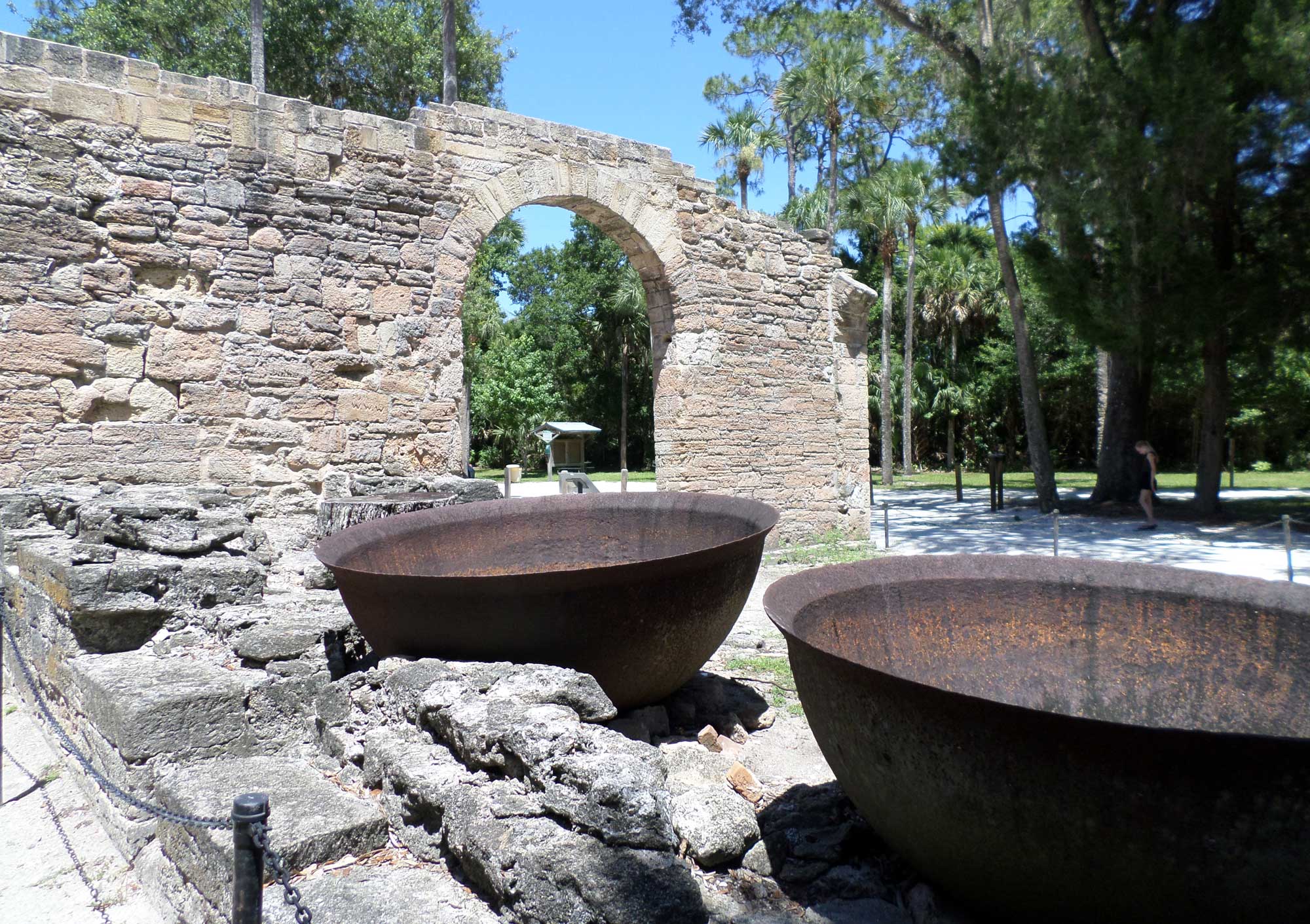 Photograph of the ruins of a sugar mill in New Smyrna Beach, Florida, dating to 1830 to 1835. The photo shows the partial wall of a stone building in the background, with two rounded kettles used for boiling sugarcane juice in the foreground. 
