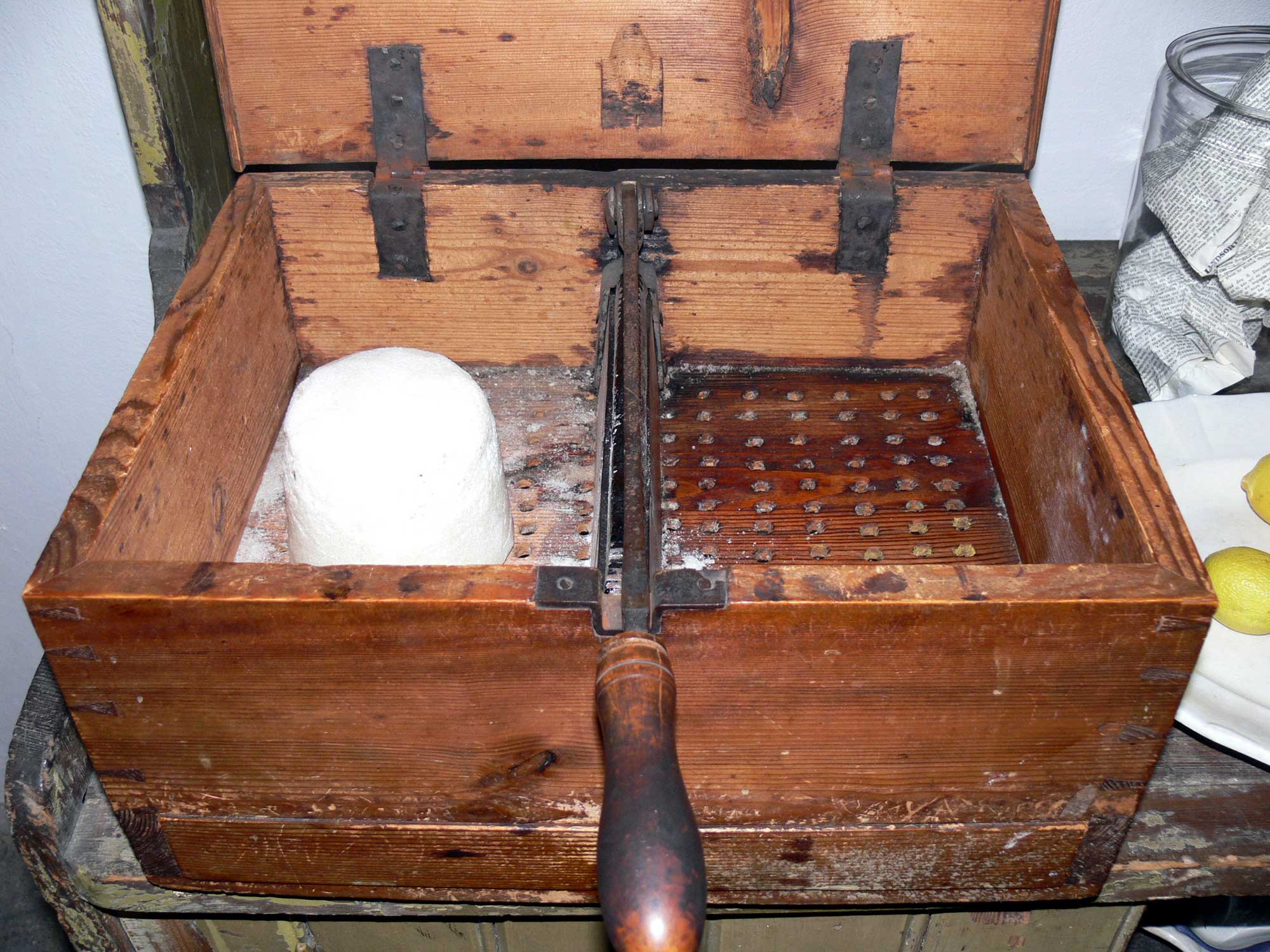 Photograph of a box used for cutting up sugar loaves. The photo shows a wooden box with a lid. The lid is open to show a knife for cutting the sugar loaf bisecting the box. The handle of the knife is sticking out of the box. Rows of holes in the bottom of the box lets sugar fall through the bottom, where it is presumably caught in a tray below.