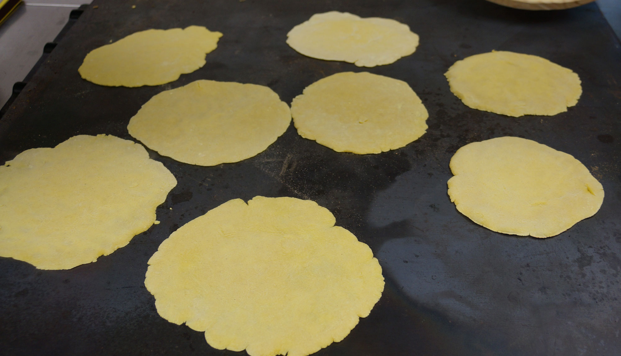 Photograph of talos cooking on a flat metal griddle. The photo shows flat, round, uncooked flatbreads made of maize flour cooking on a black metal griddle.