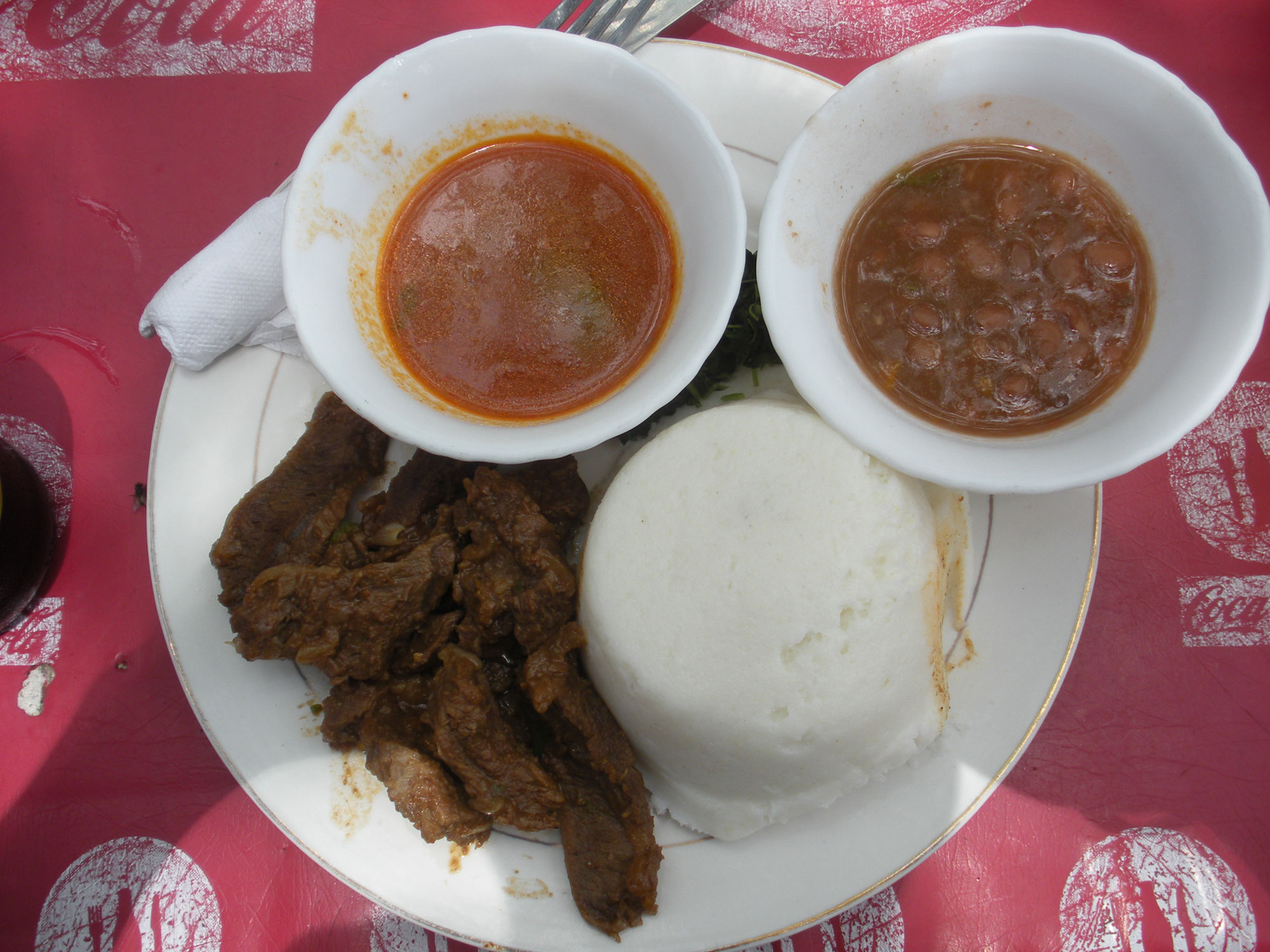 Photograph of a plate of food from Tanzania. The photo shows a white plate with pieces of beef, two bowls of sauce, and a lump of cornmeal on it.