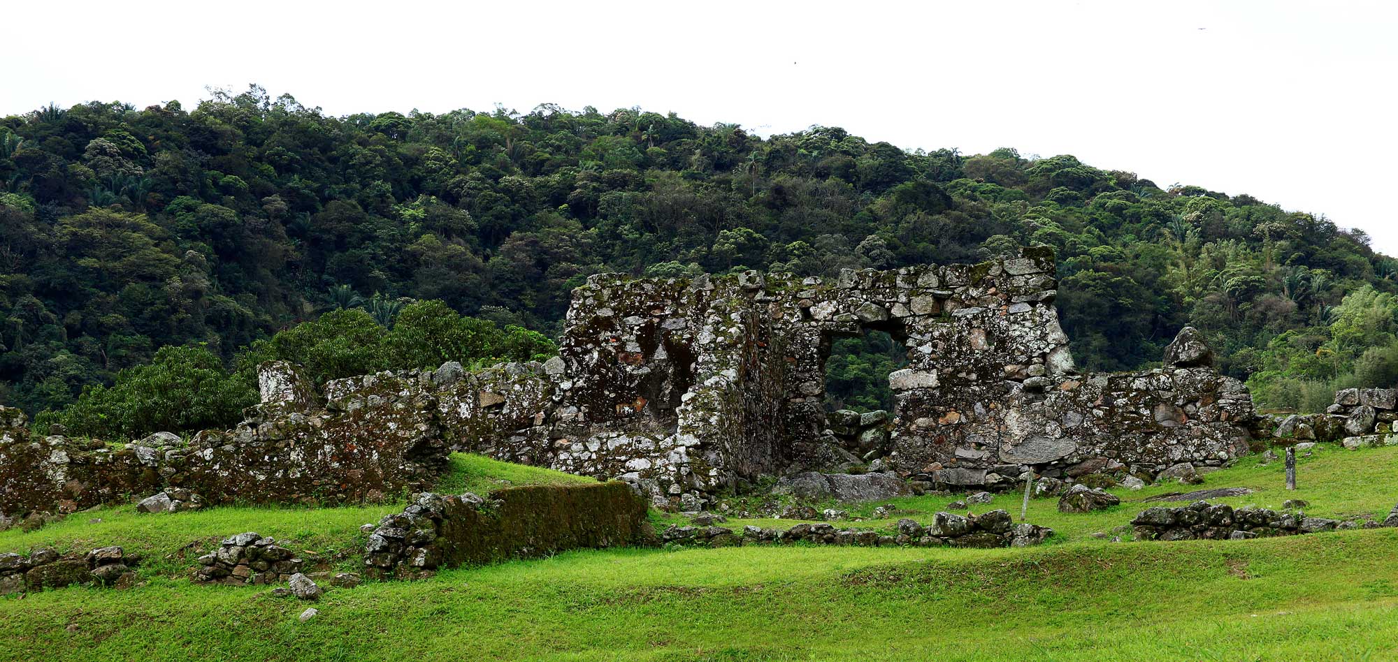 Ruins of a stone sugar mill in Brazil, from a mill first constructed in 1534. The photo shows part of a stone building with a green lawn surrounding it, and a forested hill rising behind it.