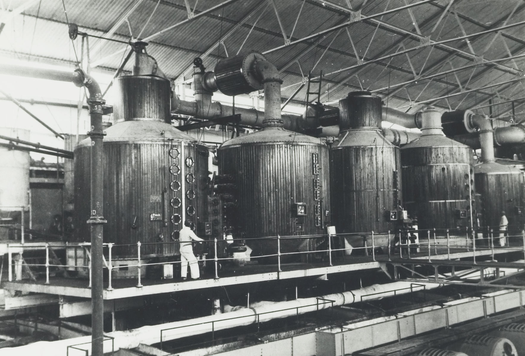 Black and white photograph of the inside of a sugar mill showing vacuum pans, Java, around 1940. The vacuum pans are a row of tall metal cylinders.
