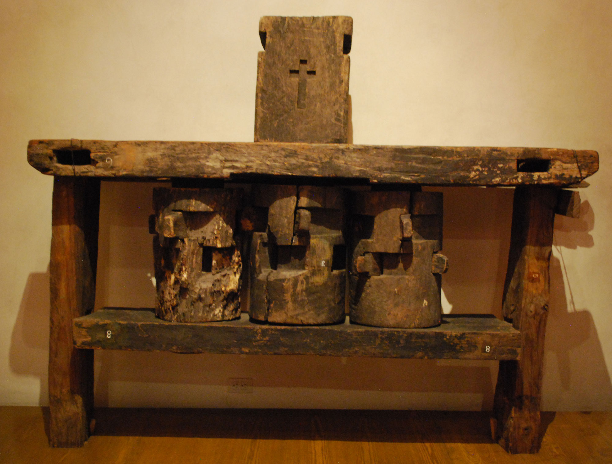 Photograph of wooden vertical sugarcane rollers on display in Mexico. The photo shows three wood rollers mounted in a wooden frame. A board with a Christian cross carved in it stands on top of the frame.