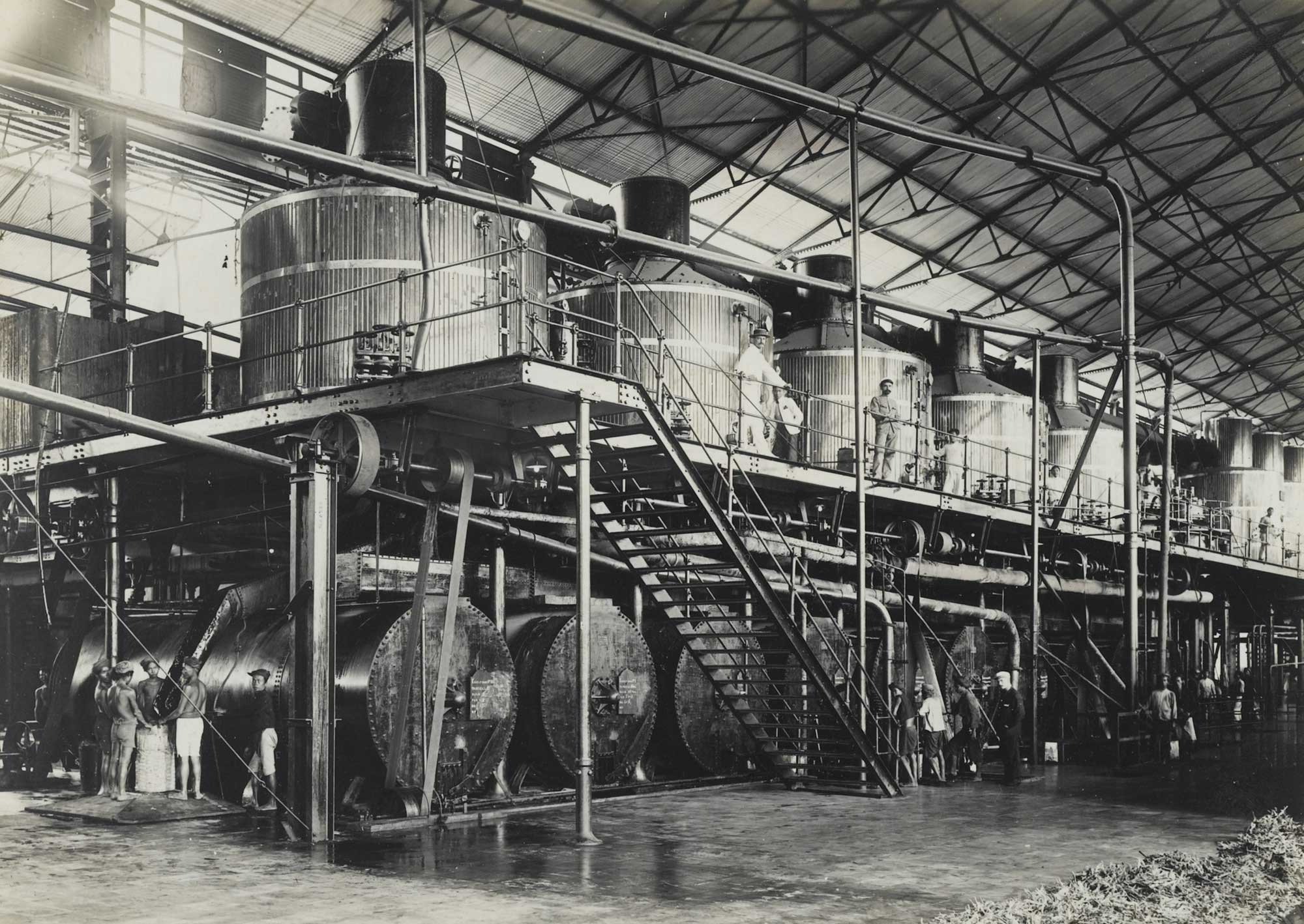 Black and white photograph of the inside of a sugar mill showing vacuum pans, Java, around 1915. The vacuum pans are a row of tall metal cylinders.