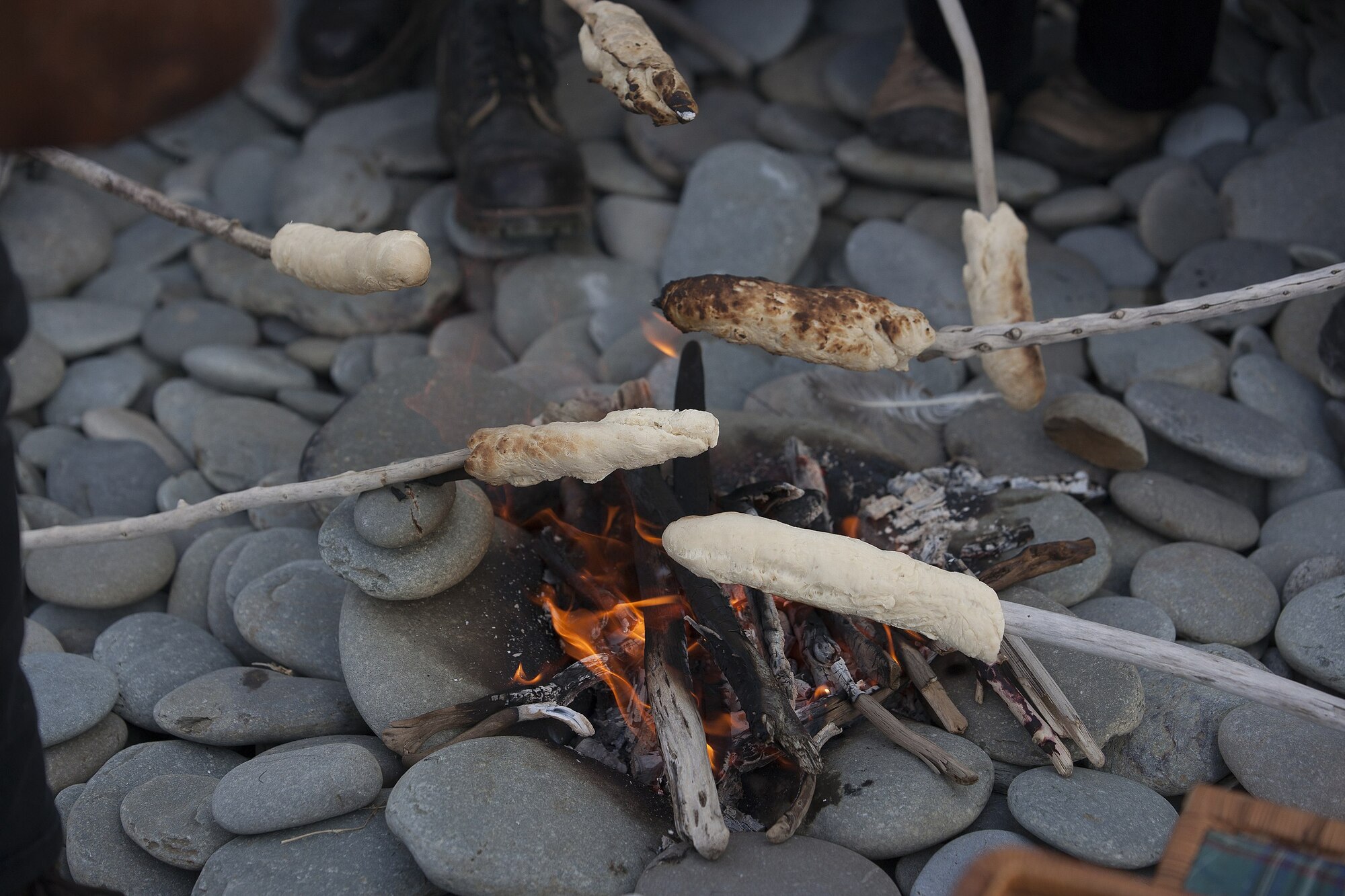 Photograph of small sticks of damper bread being cooked on wood sticks held over hot coals in a fire pit.