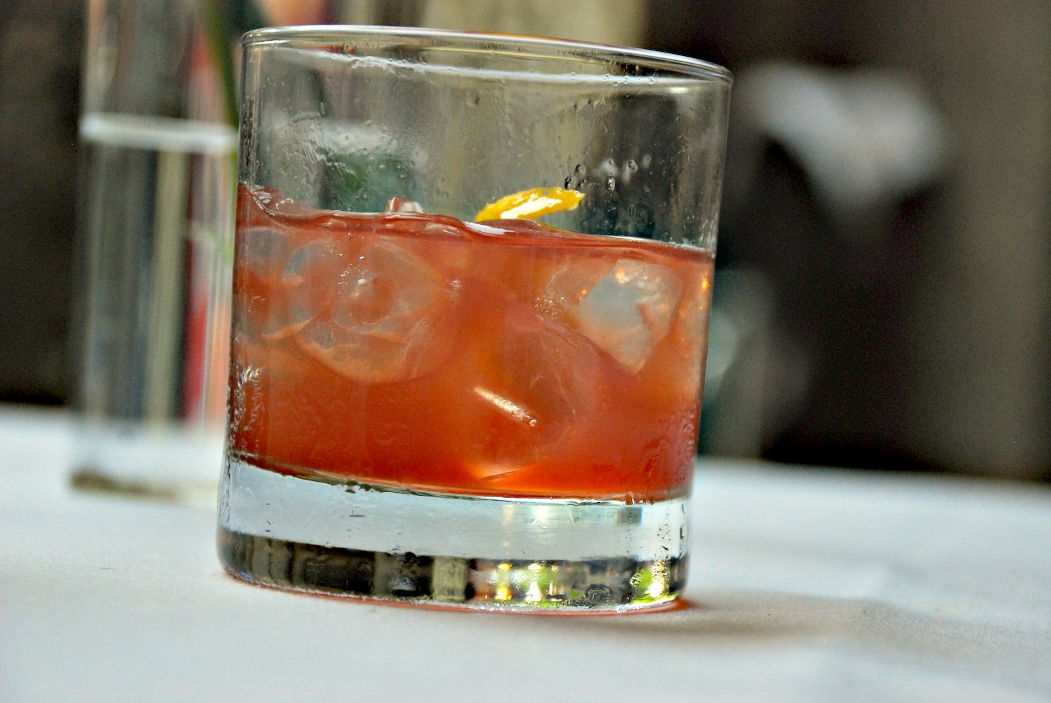 Photograph of a bourbon cocktail in a clear drinking glass. The liquid in the glass looks dark pink. The drink has ice cubes in it and perhaps a slice of lemon.