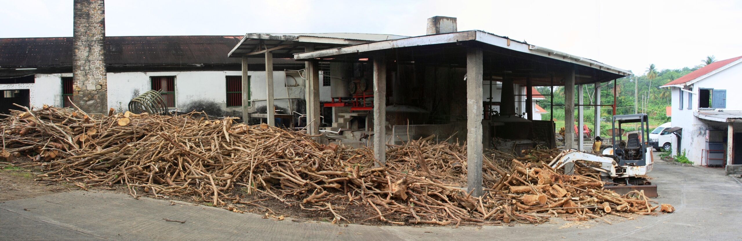 Photograph of a pile of harvested sugar cane stems next to a building where they are being processed into rum.