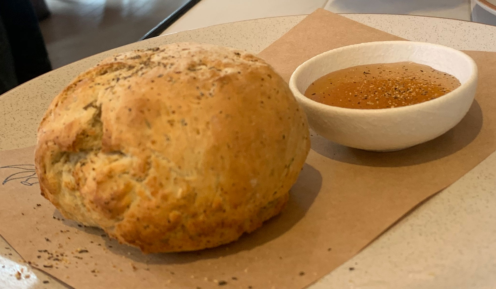 Photograph of damper, a type of bread from Australia. The photo shows a round loaf of damper bread with golden-brown crust next to a small, white bowl of oil with herbs in it. The damper bread and bowl sit on brown paper placed on a serving dish.